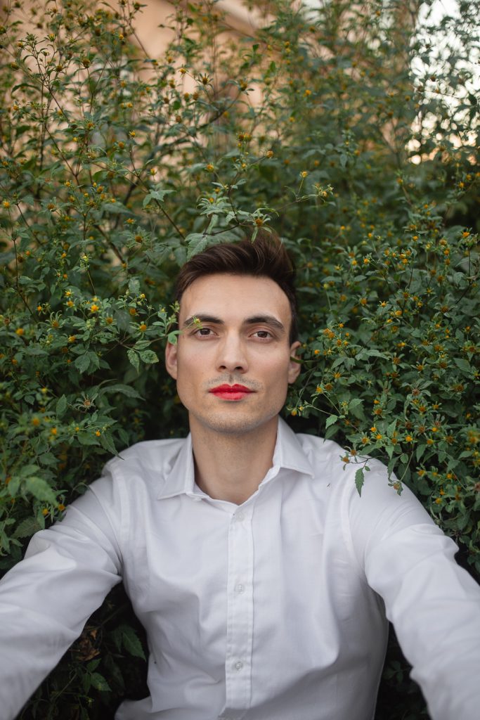 Man sitting against flowering bush wearing red lipstick