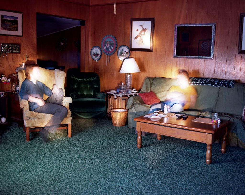 Two men sitting in a wood paneled living room with faces obscured by a bright light
